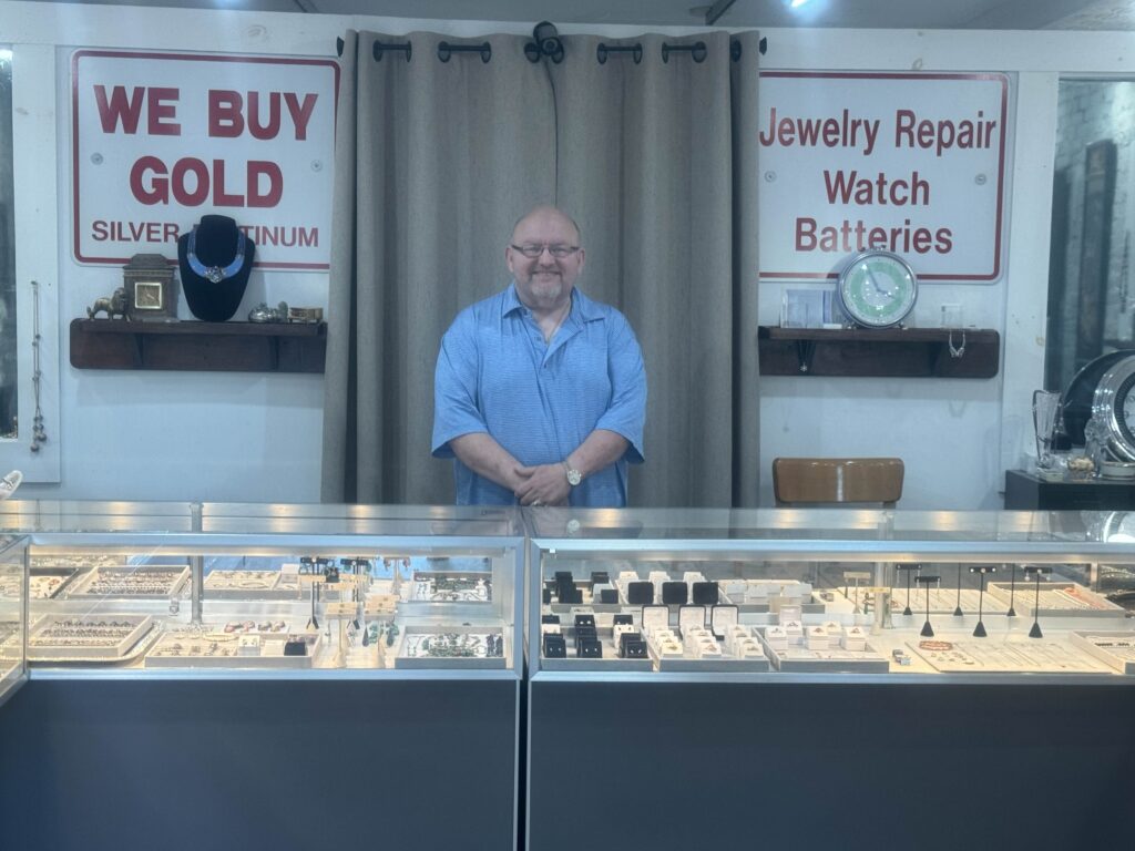 A picture of the store owner centered behind the desk with an abundance of jewelry